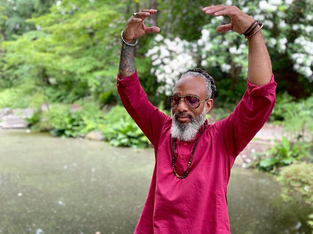 Heru TchaasAmen in a garden, arms raised in a Qigong movement